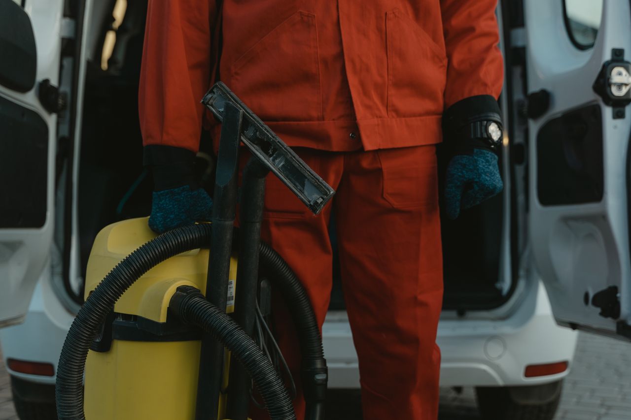 creative-01 A professional cleaner in a red uniform holding a vacuum near a van, ready for service.