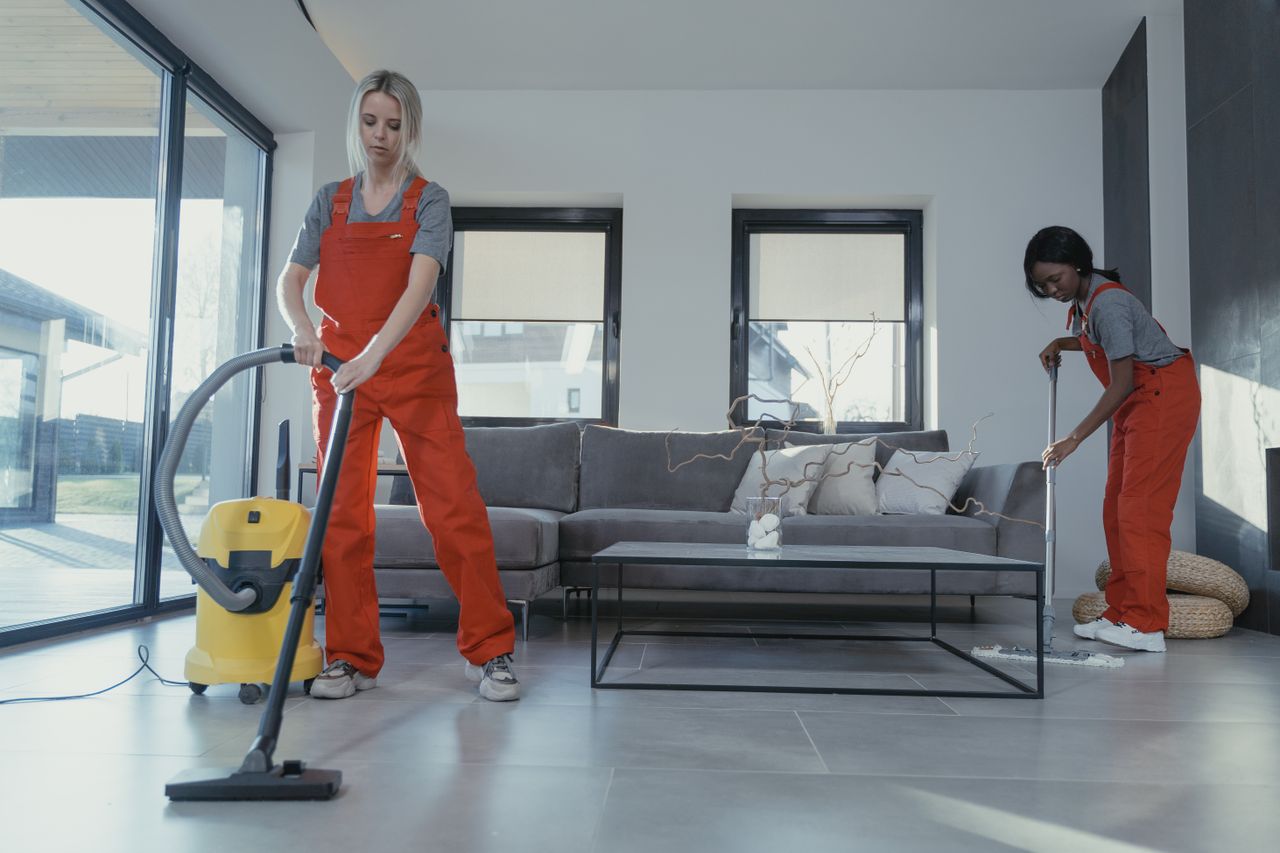 brand-03 Two women in red overalls vacuuming and sweeping a modern living room.