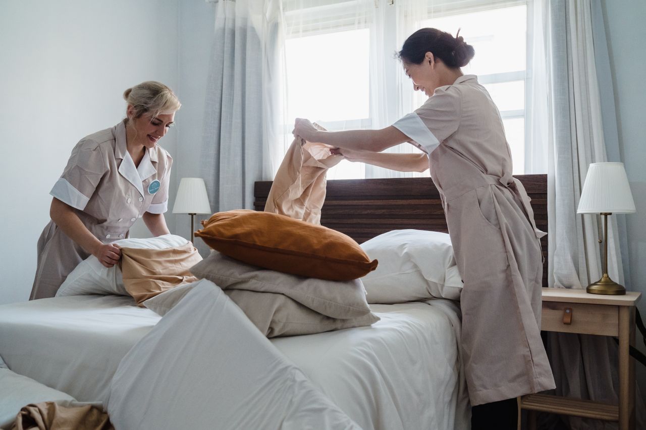brand-02 Two housekeepers in uniform fixing a hotel room bed with fresh linen and pillows.