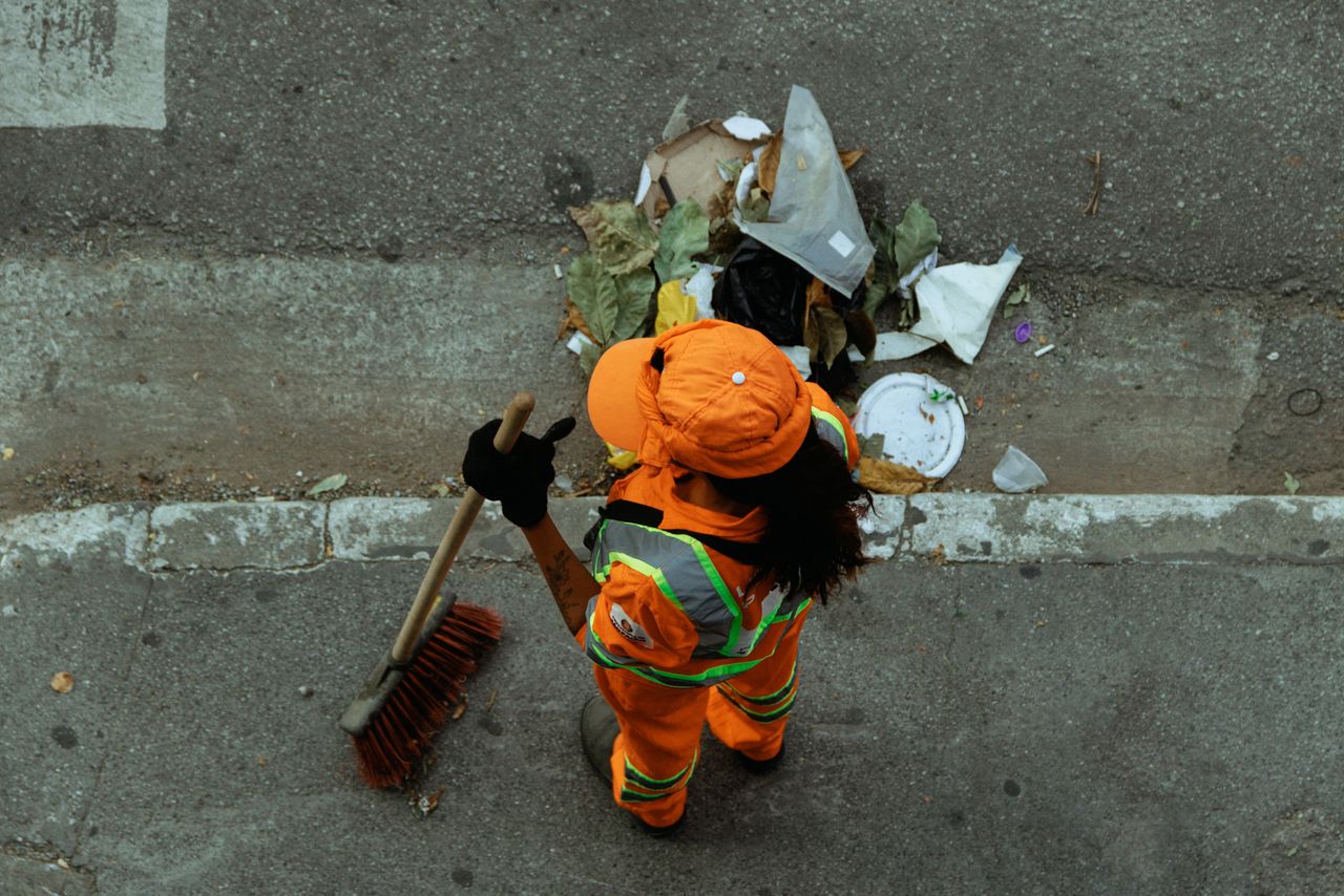 creative Top view of a street cleaner wearing orange uniform sweeping debris on a city sidewalk.